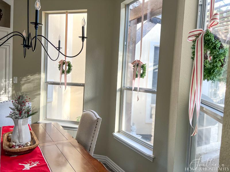 small Christmas wreaths with red and white ribbon hanging in windows, red and white stripe ribbon on wreaths, red Christmas table runner, white pitcher with Christmas greenery and red berries in dough bowl