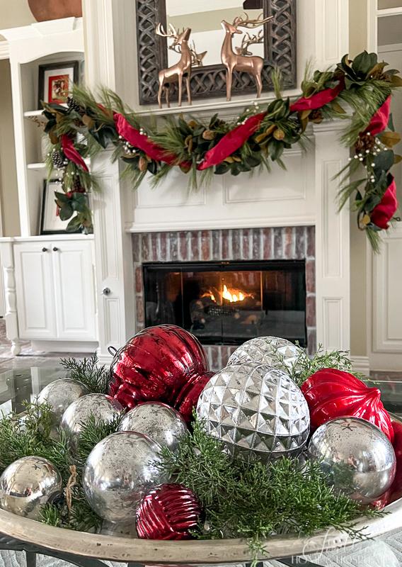 red and silver ornaments and cedar in a silver tray, in front of fireplace with magnolia and pine garland