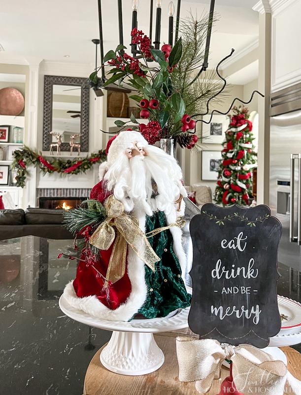 Kitchen with vintage Santa on breadboard, mercury glass vase with greenery and berries, black counters, eat drink and be merry chalkboard sign, fireplace mantel with garland, copper deer, red and white Christmas tree