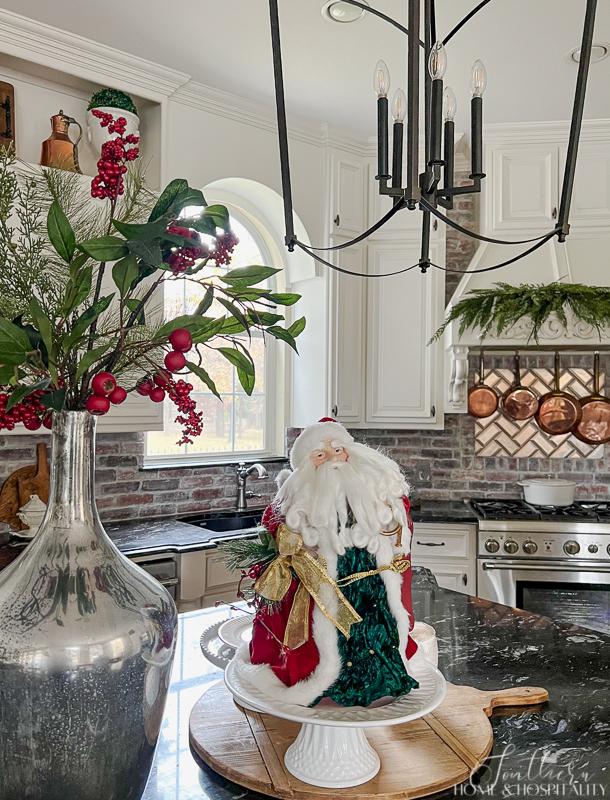 Kitchen with vintage Santa on breadboard, mercury glass vase with greenery and berries, black counters, brick backsplash, copper pots over cooktop, garland on vent hood