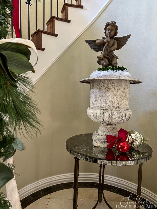 Angel on French aged urn with mercury glass red and silver ornaments beside Christmas garland on staircase