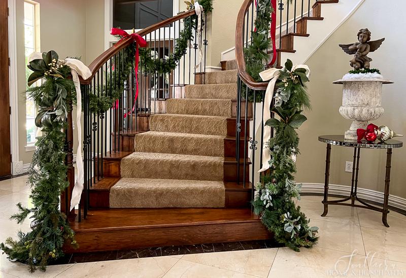 Staircase and foyer with Christmas garland of mixed greenery and ribbons draping the stair banister. Mirrored foyer table with aged French urn, angel statue, and red and white mercury glass ornaments
