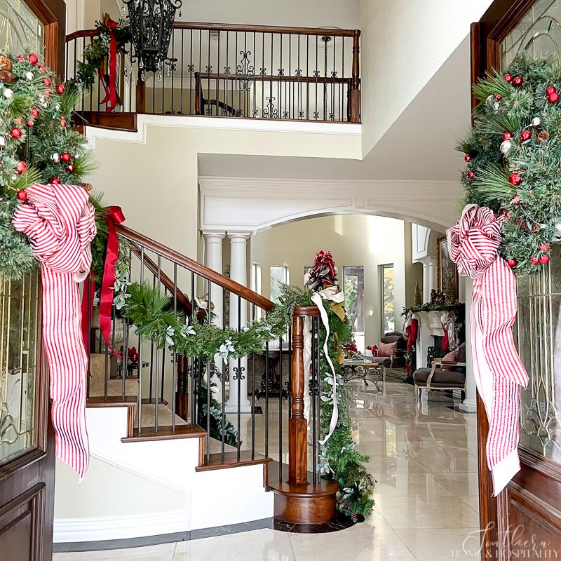 View inside double doors with Christmas wreaths of southern traditional home with garland on staircase, stockings and garland on mantel, and Christmas tree in formal living