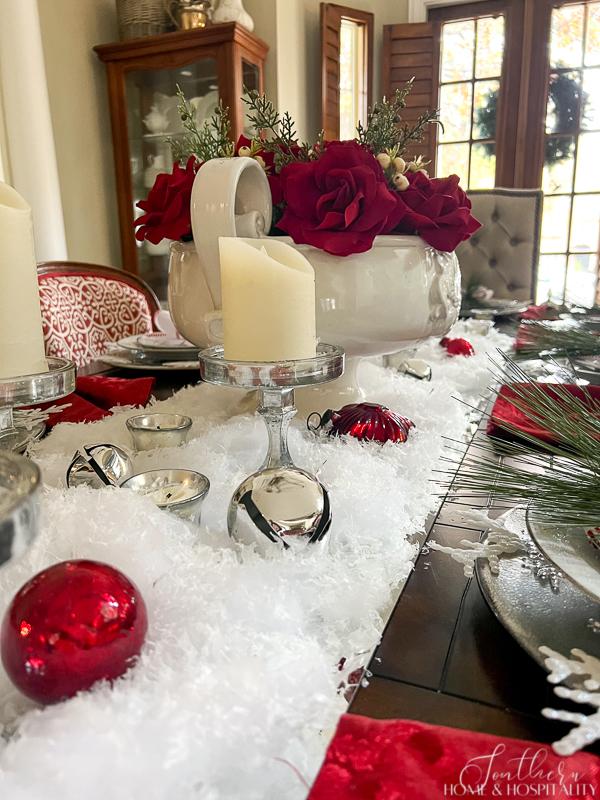 Red, silver, and white dining tablescape with artificial snow, mercury glass ornaments, jingle bells, red roses, candles