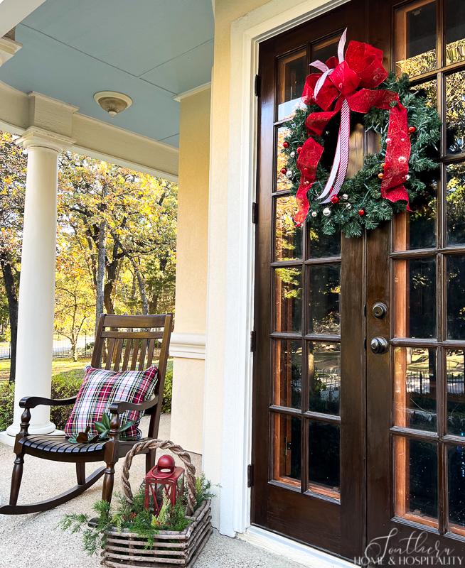 Christmas wreath hung on french doors next to rocker with plaid Christmas pillow and lantern and greenery arrangement in basket