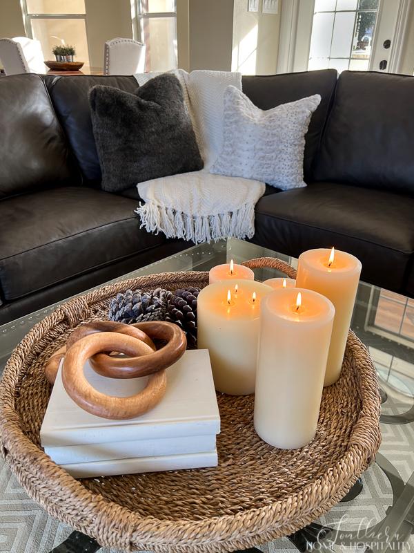 winter coffee table arrangement on wicker tray with candles, pinecones, wood decor, books, leather sectional with fur pillows, white throw blanket