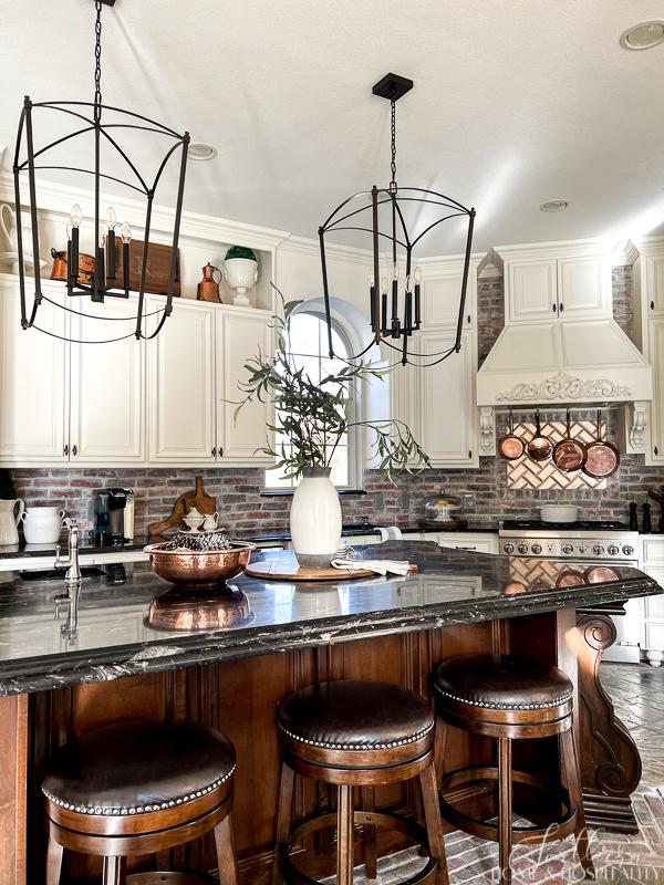 French country kitchen, brick backsplash, copper pots over range, furniture kitchen island, oversize kitchen pendants, black granite counters, white and stained cabinets, brick and slate floor