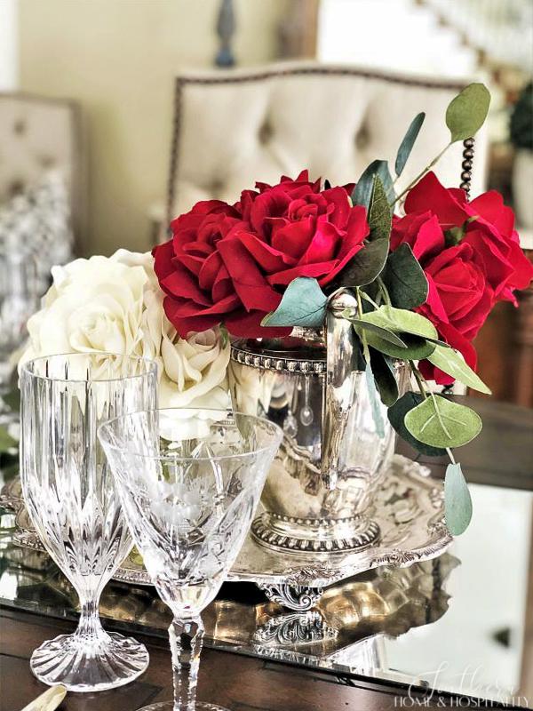 Red roses in a silver pitcher on a mirror with crystal glasses