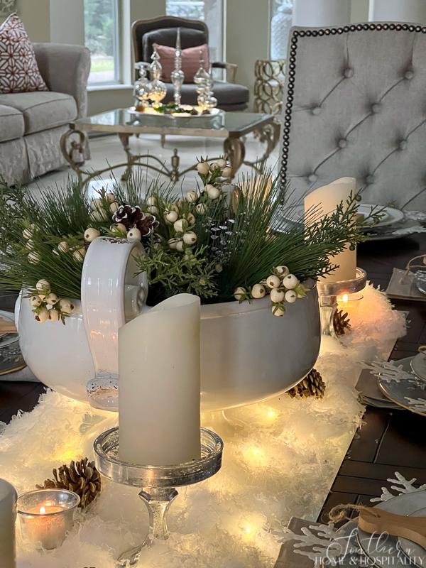 Winter table centerpiece with pine, cedar, and white berries in a pedestal bowl, white snow with fairy lights and candles, mercury glass finials