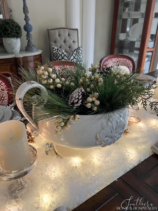 Winter tablescape with snow, candles, and winter greenery, pine and white berries in white bowl centerpiece, French sideboard, ironstone