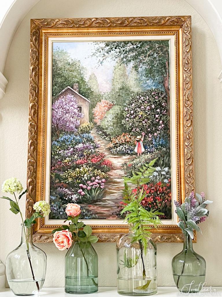 Colorful traditional painting of a garden in a gold frame over a glass vases of flowers on a fireplace mantel