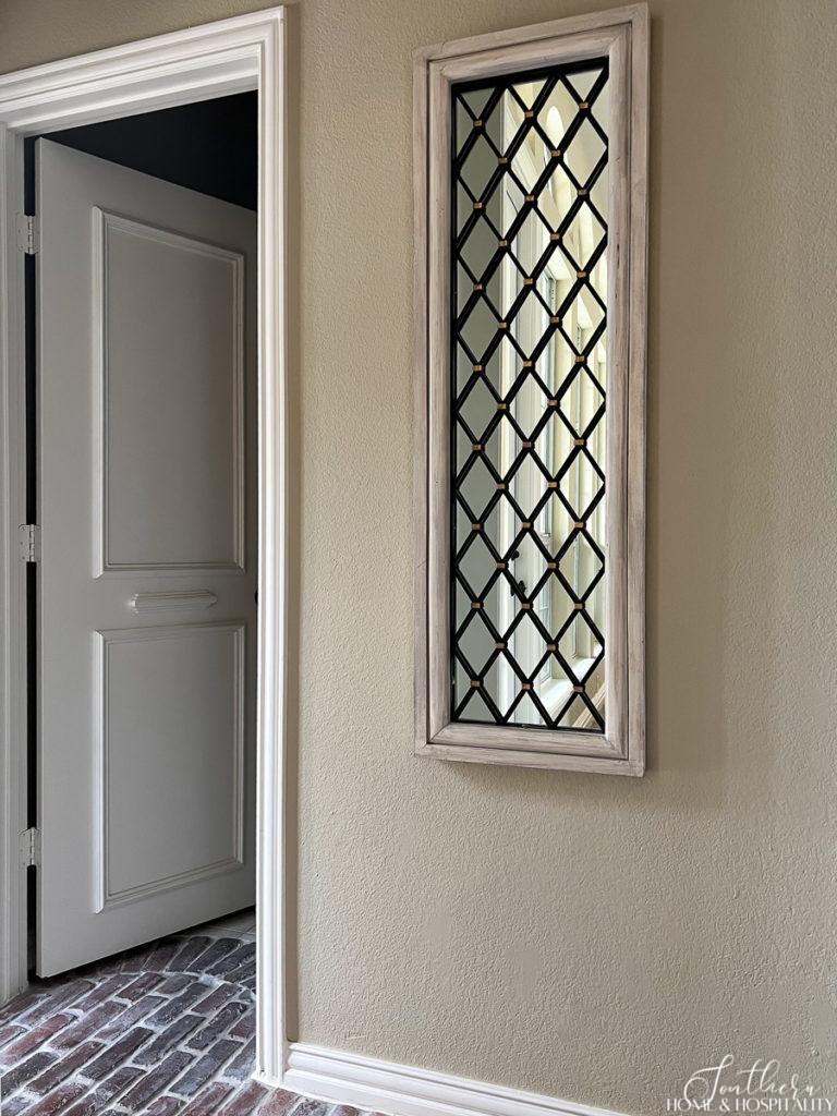 A tall narrow mirror with lattice work hung on the wall in a hallway with a brick floor