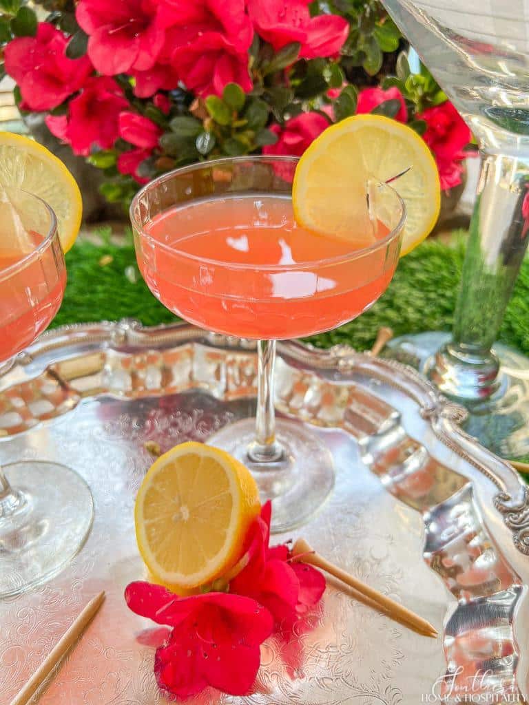 Pink Azalea cocktail in a coupe glass garnished with lemon sitting on a silver tray with golf tees and azaleas