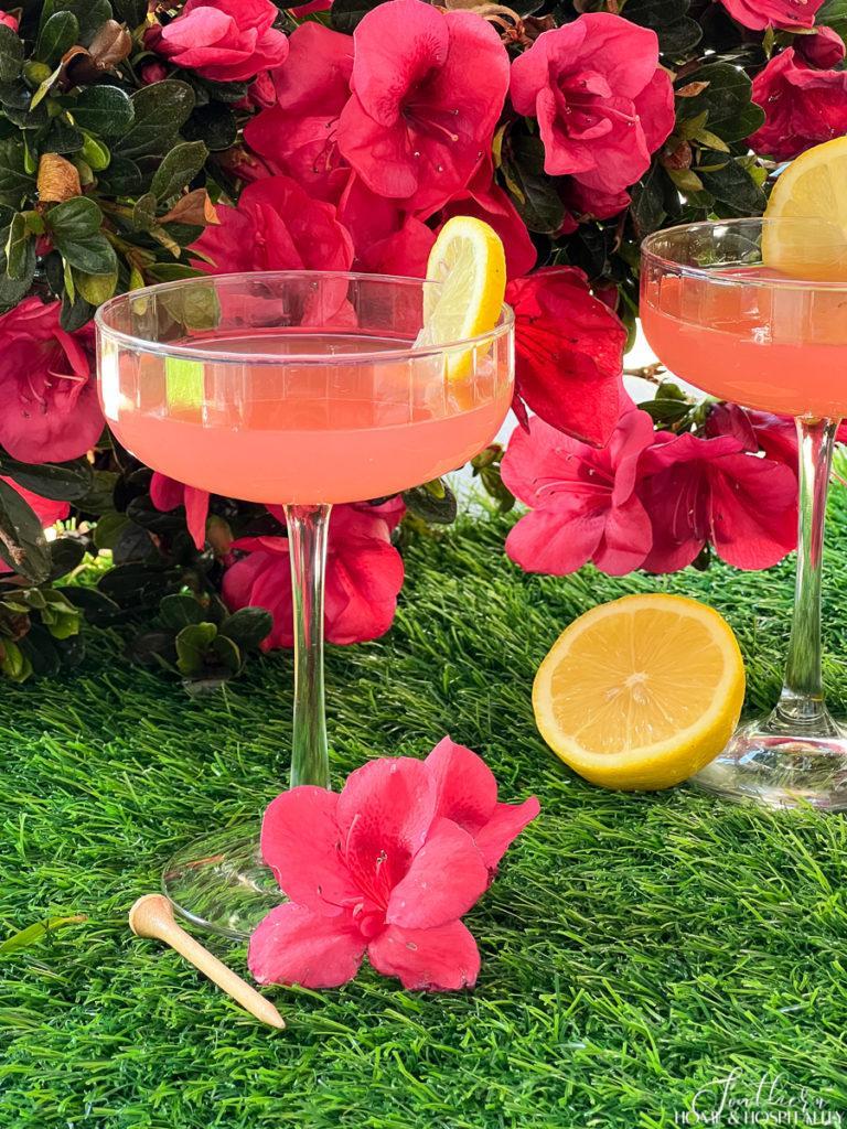 Pink Azalea cocktails in coupe glasses garnished with lemon sitting on grass with golf tees and azaleas