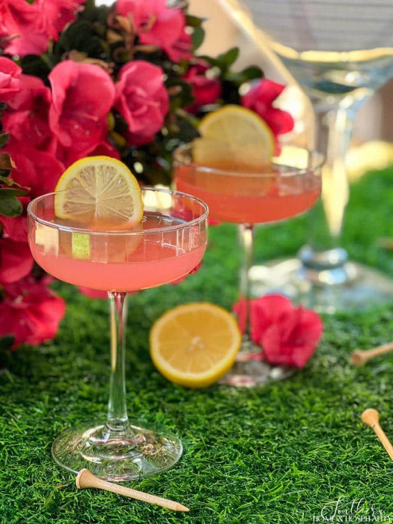 Pink Azalea Masters drink in coupe glasses garnished with lemon sitting on grass with golf tees and azaleas