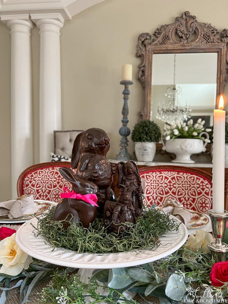 Faux chocolate bunnies on green spanish moss on a white cake pedestal in an Easter centerpiece