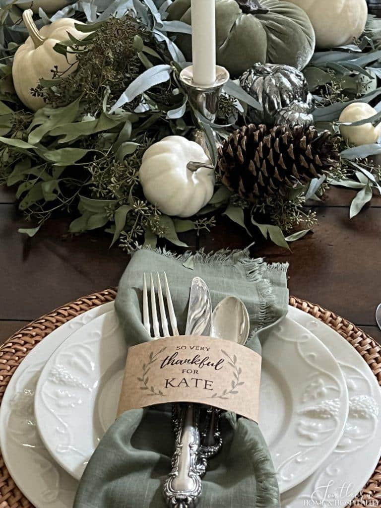Thanksgiving place setting with silver utensils and sage green napkin wrapped in a custom name placecard set on white ironstone dishes