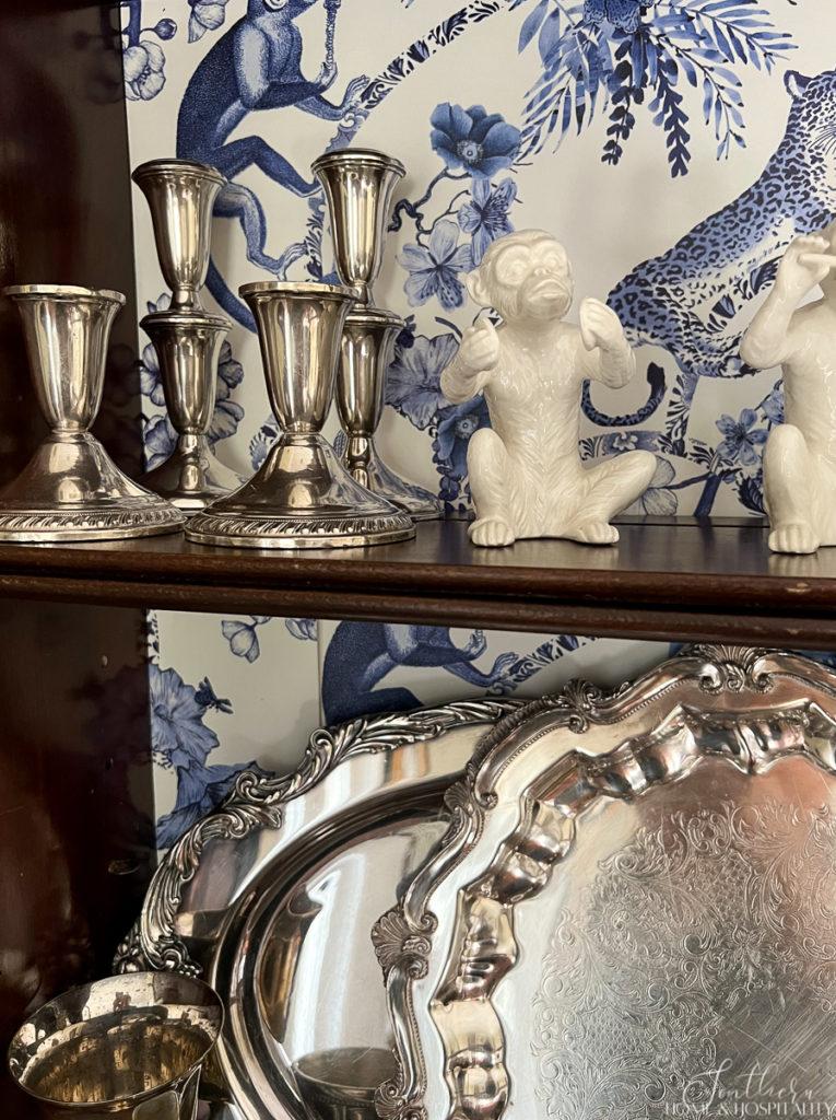 Vintage silver candlesticks and trays in a wood cabinet with blue and white chinoiserie wallpaper back