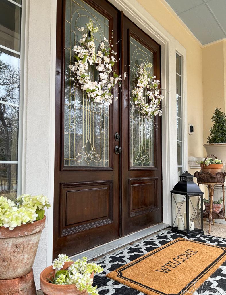 Dogwood and cherry blossom spring wreaths on double wood front doors on a southern porch decorated for spring with welcome mat and spring flowers in garden pots
