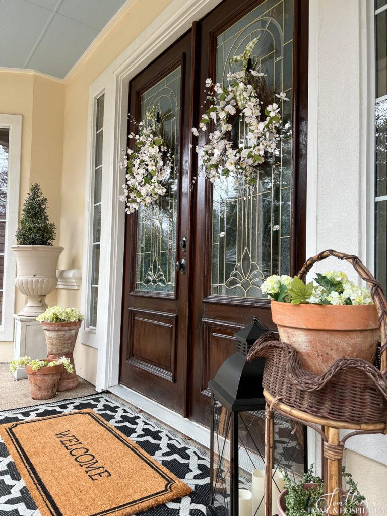 White flower spring wreaths hanging on double wood front doors on a southern porch with welcome mat and spring flowers in garden pots