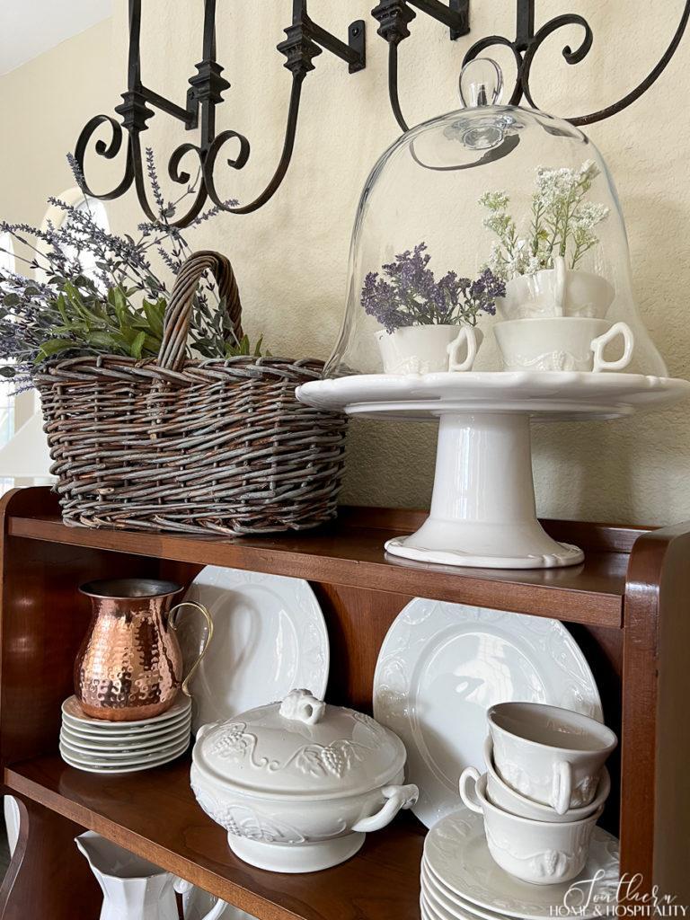 Vintage hutch with ironstone dishes and decorated for spring with purple and white flowers in teacups under a cloche and lavender in a basket