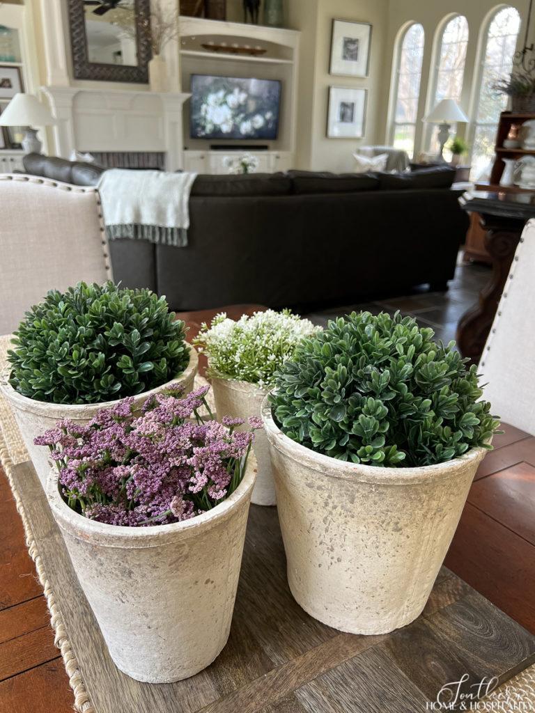 Spring kitchen table centerpiece in a French country kitchen with rustic garden pots, boxwood, and purple and white flowers on a charcuterie board with natural table runner