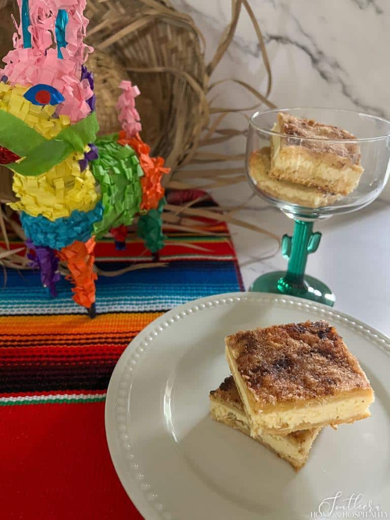 sopapilla cheesecake bars served on white plate and in a cactus margarita glass, on a mexican table runner with a mini pinata