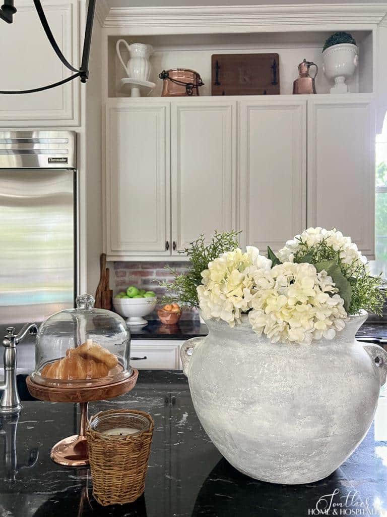 White hydrangeas in rustic garden pot, wicker candle, and copper cloche on kitchen counter