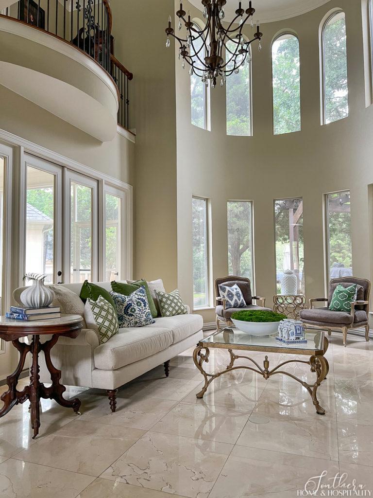 Traditional two story living room with white linen sofa, blue and green throw pillows, Bergere chairs, and antique French marble top table