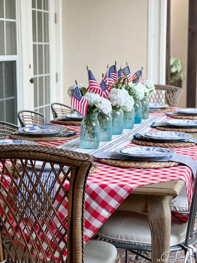 Elegant patriotic tablescape set on French country dining table with red check, seersucker, mason jars, flags, and white flowers