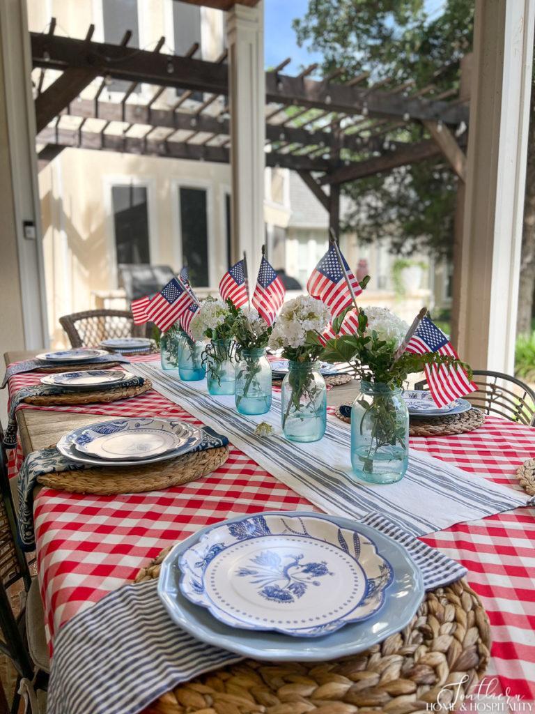 Fourth of July centerpiece with blue mason jars, white flowers, and flags, blue and white dishes, seersucker napkin