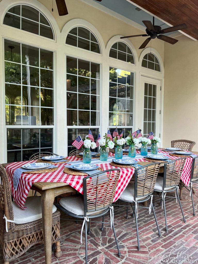 Elegant patriotic tablescape set on French country dining table on a brick patio with red check, seersucker, mason jars, flags, and white flowers