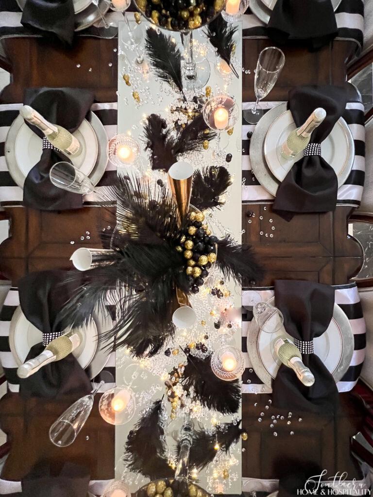 Black, gold, and white New Years tablescape with feathers, candles, mirror, and bow napkins