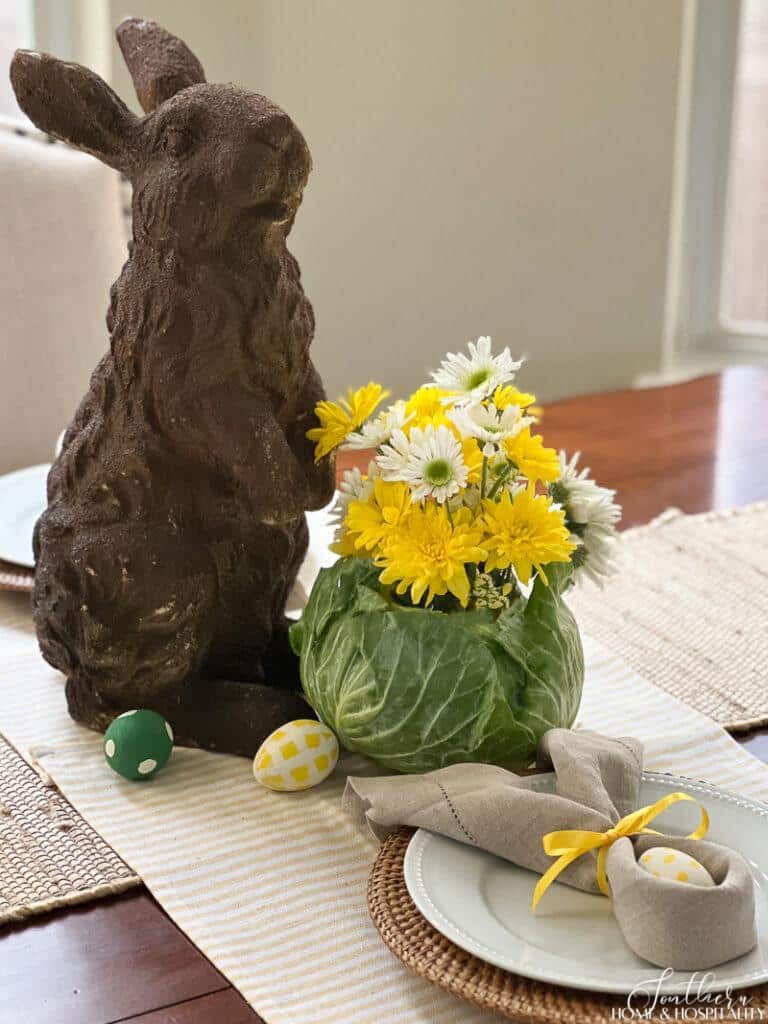 Easter table centerpiece with rabbit garden statue and yellow and white fresh flowers in a cabbage vase