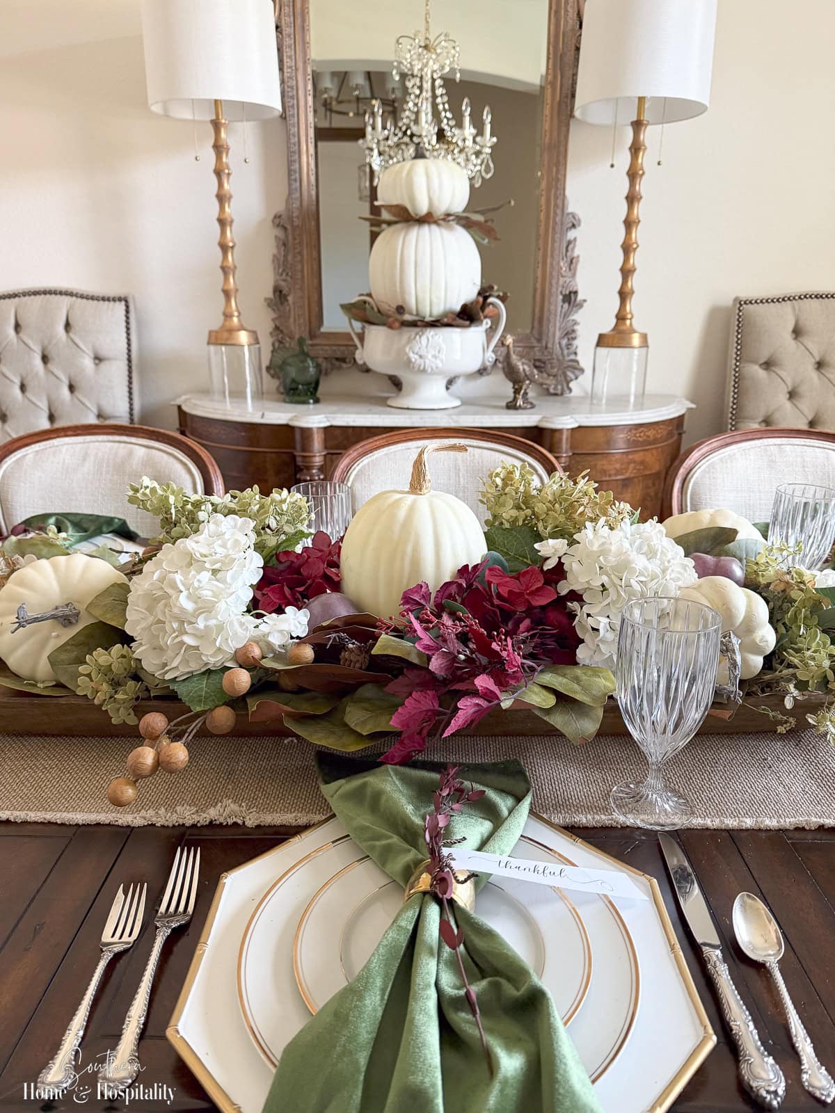 Burgundy and green Thanksgiving centerpiece in a long wooden dough bowl filled with hydrangeas, magnolia leaves, and white pumpkins with green velvet napkins.