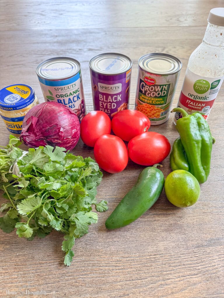 Flat lay of cowboy caviar ingredients including canned beans, bell peppers, jalapeno, garlic, corn, tomatoes, onion, cilantro, and Italian dressing.