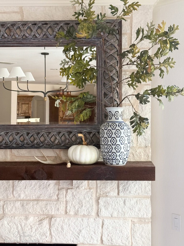 Blue and white vase with muted green fall branches, a white pumpkin, and a deer antler decorating a fall mantle.