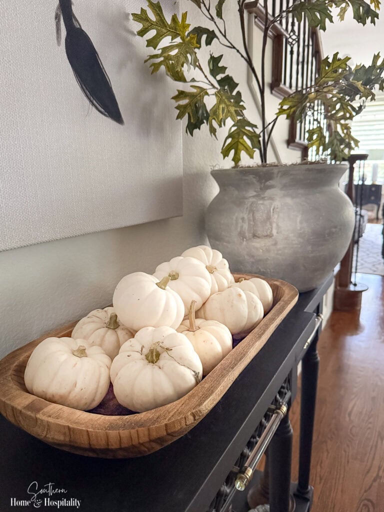 Dough bowl filled with white pumpkins on a black entry table.