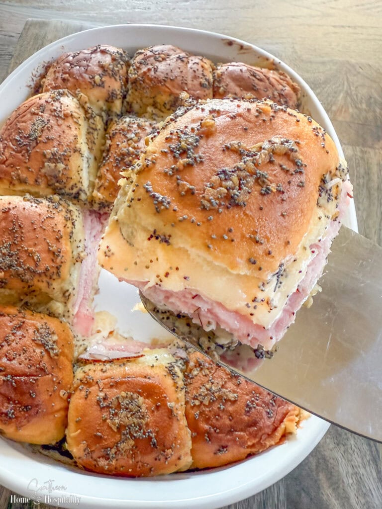 Hot ham and cheese slider on a spatula lifted from pan with melted Swiss cheese and buttery poppyseed glaze.
