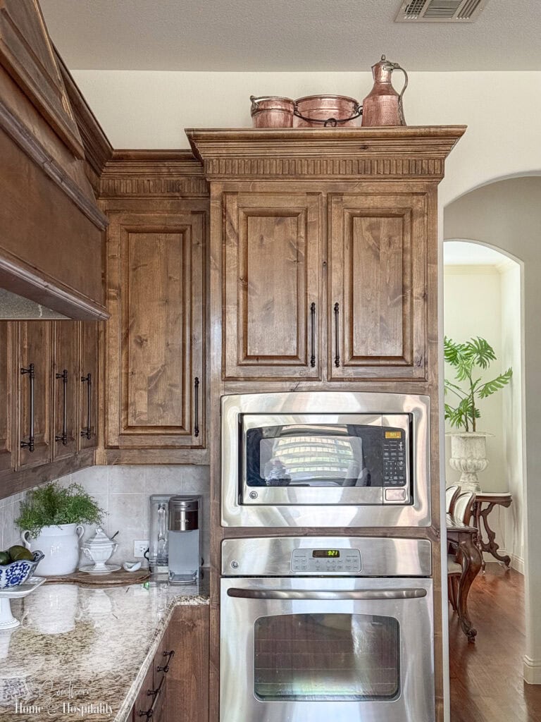 Grouping of copper pots and pitchers arranged above kitchen cabinets, warm wood tones and brown granite counters below, minimal decor for a classic kitchen