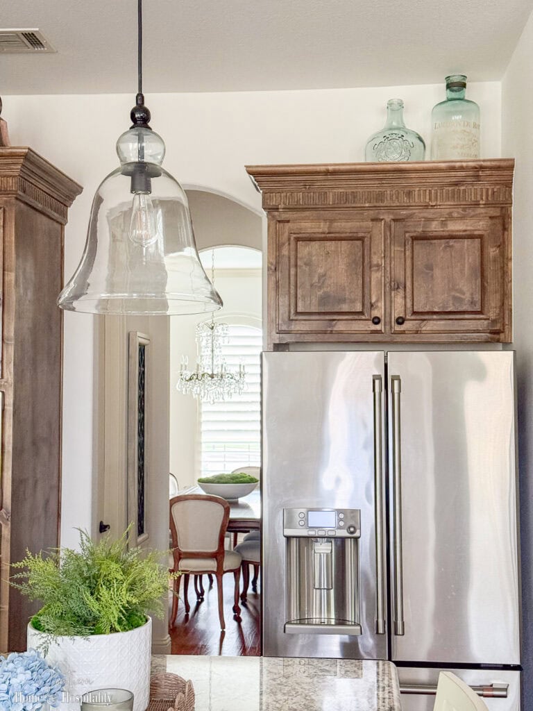 Clear glass wine jug and tall bottle displayed above upper kitchen cabinets, simple minimal decor that adds light reflection and keeps the space airy