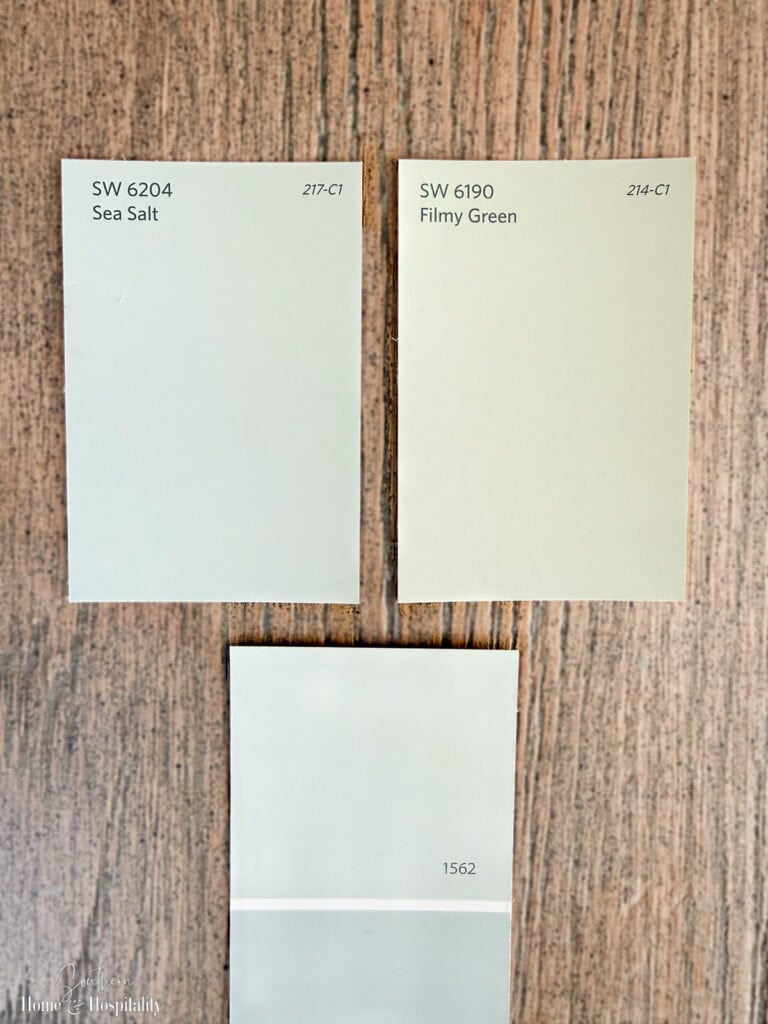 Muted sage green paint samples including Sea Salt, Filmy Green, and Healing Aloe displayed on a wood surface.