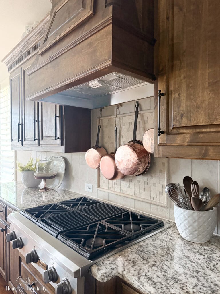 Clean kitchen stovetop and vent hood with hanging copper pots in a warm wood cabinet kitchen.