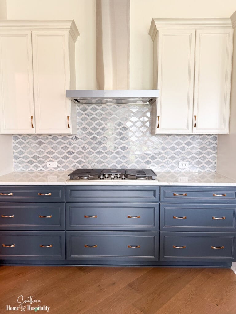 Cooktop wall with patterned backsplash, white cabinetry, blue lower cabinets, quartz countertops, and brass hardware