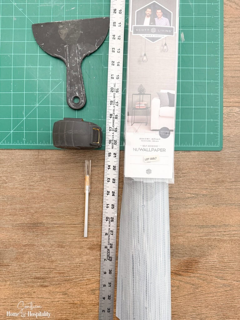 Supplies for wallpapering shelf backs including peel-and-stick wallpaper, measuring tape, ruler, X-Acto knife, smoothing tool, and cutting mat.