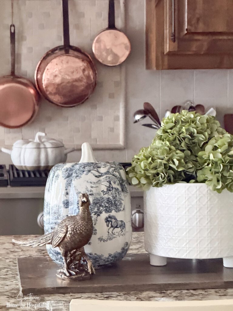 Fall kitchen island vignette with blue and white pumpkin, green hydrangeas, white planter, and brass pheasant
