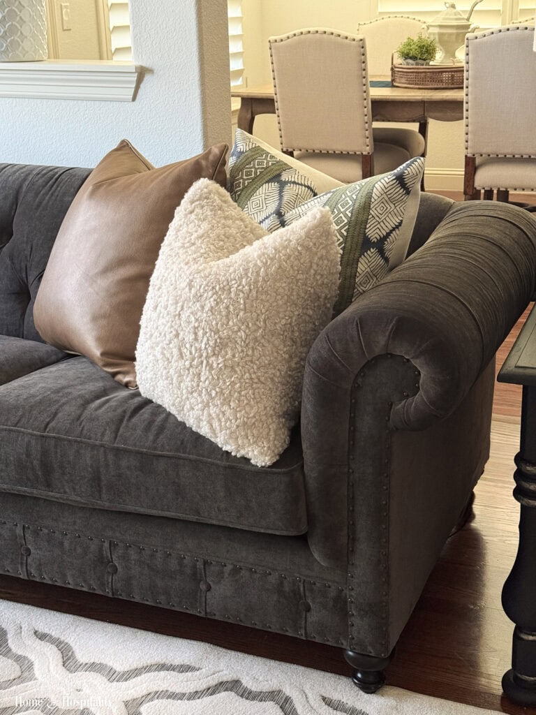 Cream bouclé, cognac leather, and navy and olive patterned pillows layered on a charcoal velvet sofa