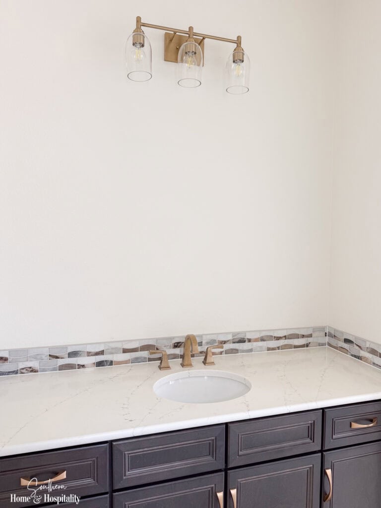 New home bathroom vanity with white countertop, brass fixtures, and wall left open for a decorative mirror