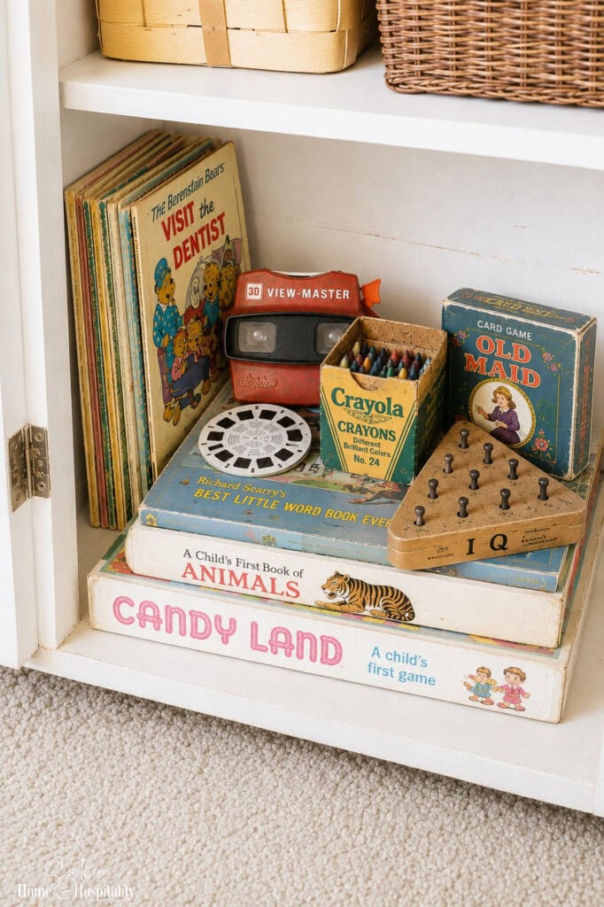 Cabinet filled with vintage children’s toys and games including Candy Land, Old Maid, a View-Master, crayons, books, and a wooden peg game.