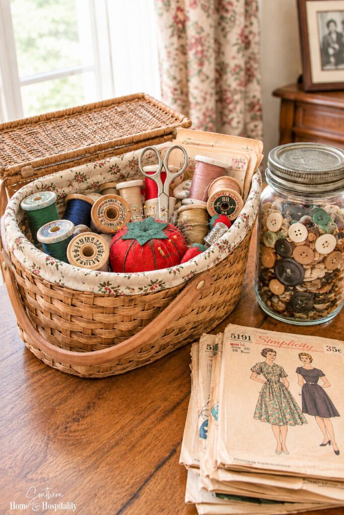 Vintage sewing basket filled with thread, scissors, a tomato pincushion, sewing patterns, and a glass jar of saved buttons.